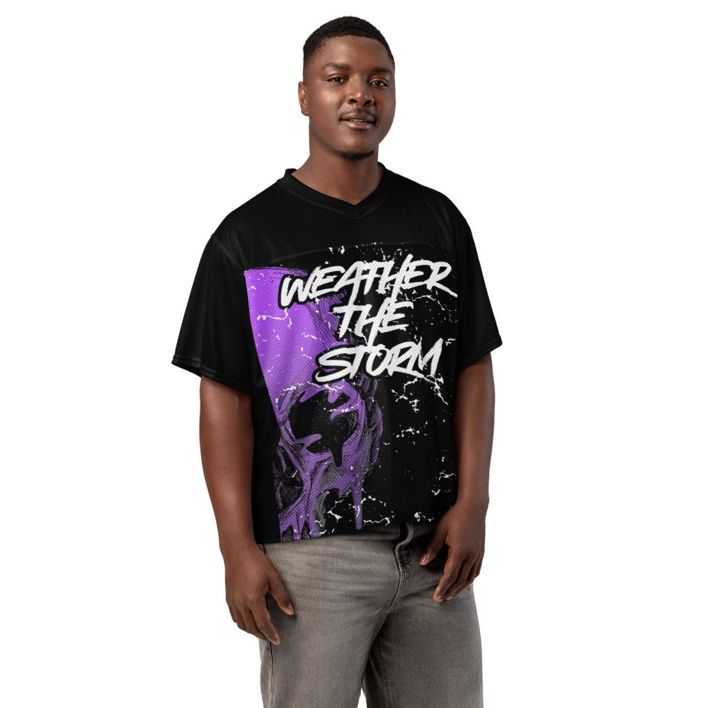 A man wearing a black V-neck t-shirt with a purple and white graphic that reads 'WEATHER THE STORM' on the front, paired with gray jeans, set against a plain white background.