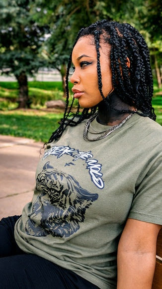 A young black woman with black braids sits outdoors at bench in Manitou Springs, Colorado in September. She is wearing the green graphic t-shirt with a tiger print and that says via  text 'BREAK THE MOLD' designed by Fittwave Fitness Inc. a 501c3 non-profit based in Colorado Springs, Colorado. 