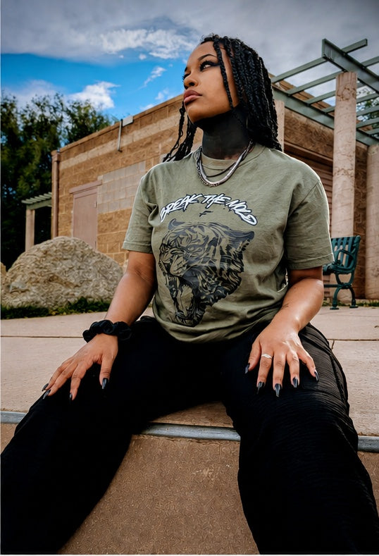 A young black woman with black braids sits outdoors at skatepark half pipe in Manitou Springs, Colorado in September. She is wearing the green graphic t-shirt with a tiger print and that says via  text 'BREAK THE MOLD' designed by Fittwave Fitness Inc. a 501c3 non-profit based in Colorado Springs, Colorado. 