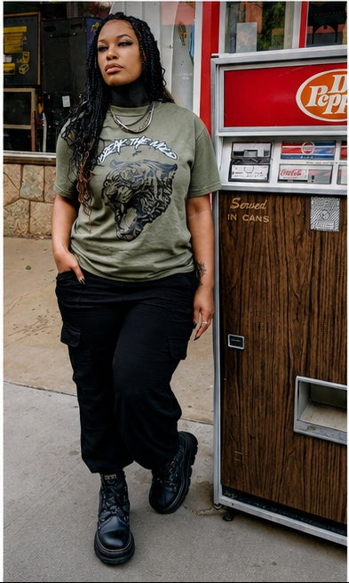 A young black woman with black braids stands next to a vintage arcade vending Dr. Pepper machine in Manitou Springs, Colorado in September. She is wearing the green graphic t-shirt with a tiger print and that says via  text 'BREAK THE MOLD' designed by Fittwave Fitness Inc. a 501c3 non-profit based in Colorado Springs, Colorado. 